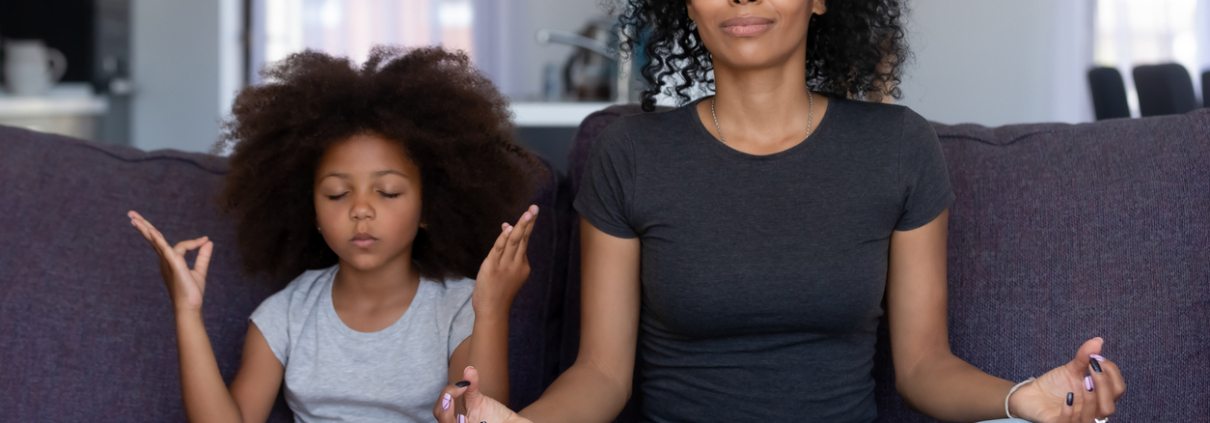 Mindful african mom with cute funny kid daughter doing yoga exercise at home, calm black mother and mixed race little girl sitting in lotus pose on couch together, mum teaching child to meditate