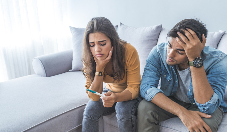 Sad stressed couple with pregnancy test sitting at home. Upset woman is looking in pregnancy test. Frustrated man is sitting next to her.  Worried about the serious consequences