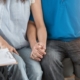 A closeup image of a man and woman seated and holding hands with a doctor writing on a clipboard beside them.