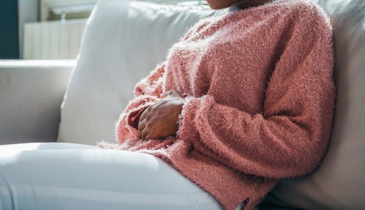 A woman in a sweater sits on her sofa with both hands on her abdomen