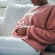 A woman in a sweater sits on her sofa with both hands on her abdomen