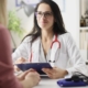 A female doctor holding a clipboard sits across from a female patient at the doctor’s office.