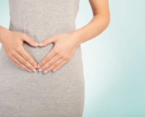A closeup image of a woman’s hands in a heart shape against her abdomen.