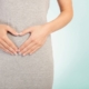 A closeup image of a woman’s hands in a heart shape against her abdomen.