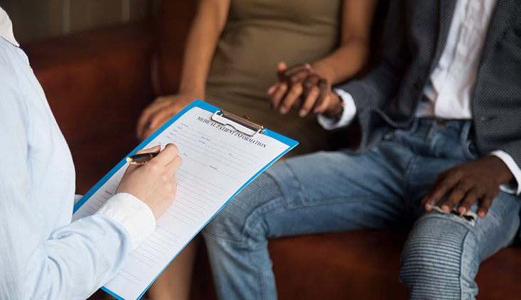 A closeup image of a couple holding hands as they sit across from a doctor holding a clipboard.