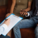 A closeup image of a couple holding hands as they sit across from a doctor holding a clipboard.