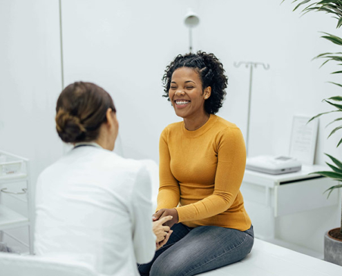 A smiling female patient sits on an exam table across from a female doctor discussing fertility options.