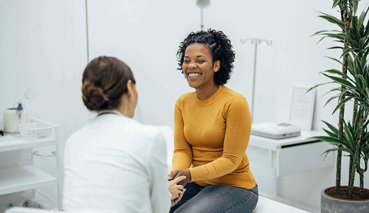 A smiling female patient sits on an exam table across from a female doctor discussing fertility options.