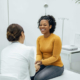 A smiling female patient sits on an exam table across from a female doctor discussing fertility options.