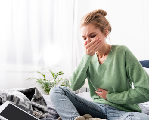 A woman sitting on her bed holding her stomach, appearing nauseous. She has a smartphone and laptop nearby.