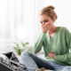 A woman sitting on her bed holding her stomach, appearing nauseous. She has a smartphone and laptop nearby.