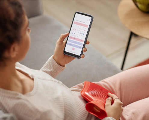 A woman sits on her sofa with a hot water bottle on her abdomen for menstrual cramp relief. She looks at a period tracker app on her smartphone.