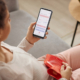 A woman sits on her sofa with a hot water bottle on her abdomen for menstrual cramp relief. She looks at a period tracker app on her smartphone.