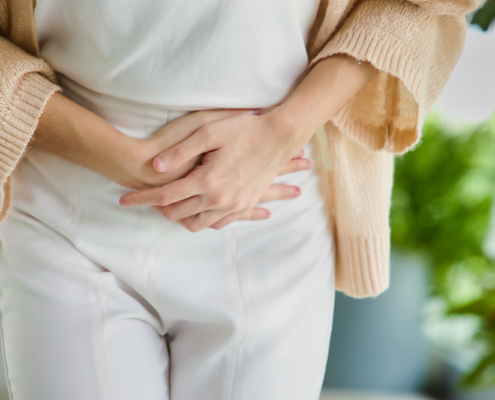 An Asian woman in a white shirt holds her stomach with both hands, appearing to experience abdominal discomfort or pain.