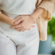 An Asian woman in a white shirt holds her stomach with both hands, appearing to experience abdominal discomfort or pain.